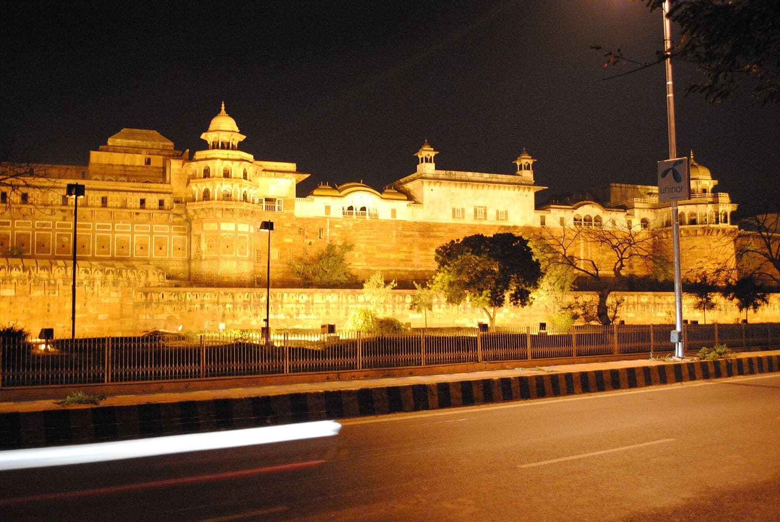 Agra Fort’s red sandstone walls lit up gold at night, with a road and streetlights in the foreground