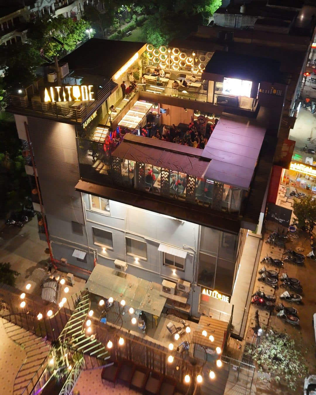 Altitude Rooftop Lounge from the air at night: the lit ALTITUDE sign on the corner of Jaipur House, the terraces glowing on the top floor and MG Road 2 below