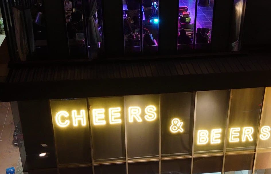 The Cheers & Beers neon sign glowing on the glass front of its first-floor bar at night, with lit windows on the floor above