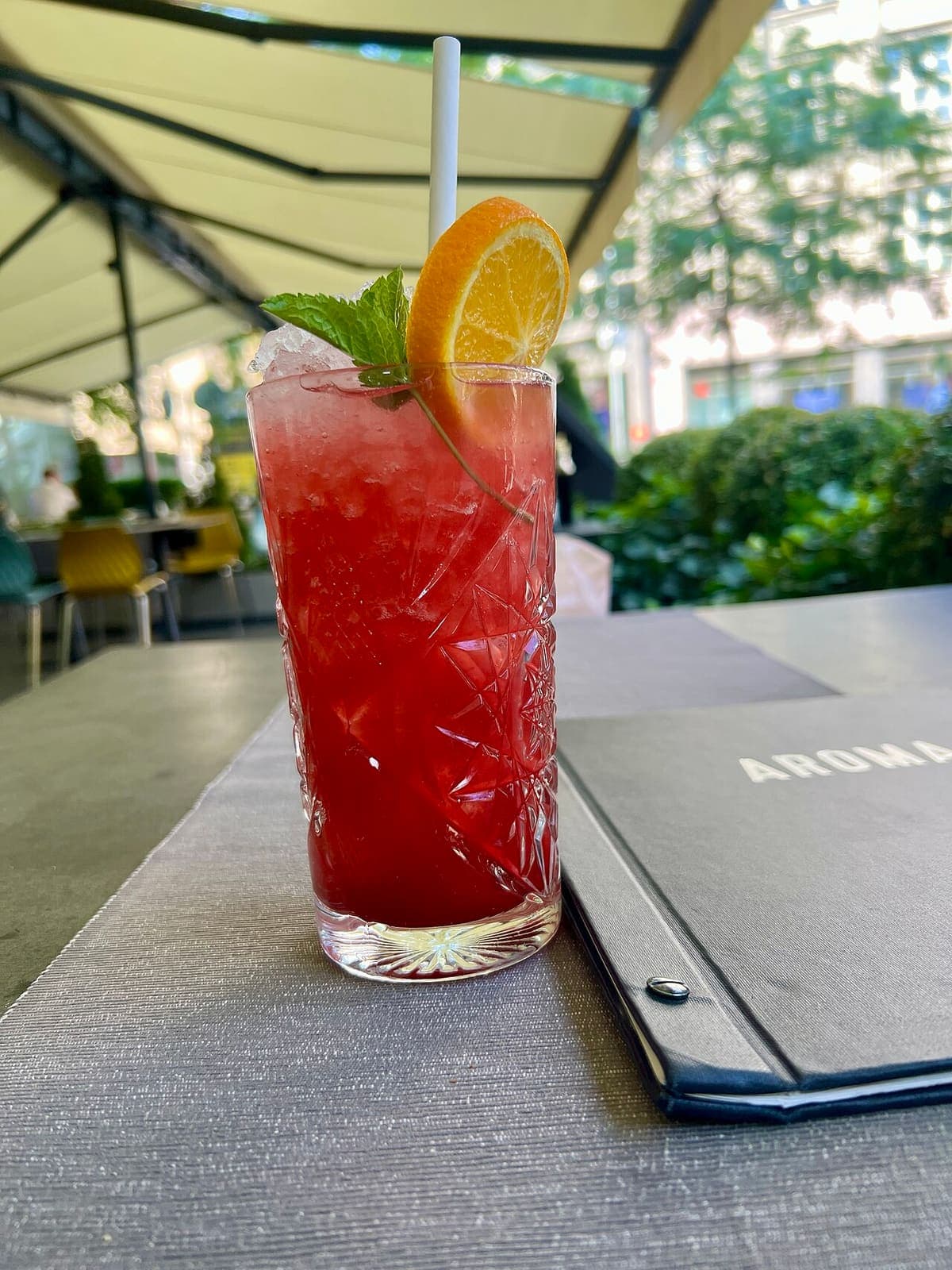 A tall red fruit mocktail with orange and mint, on an outdoor café table