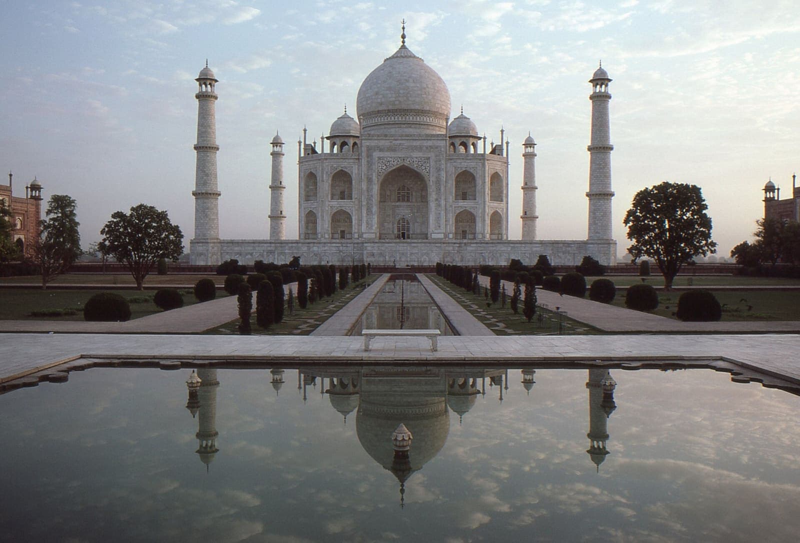 The raised marble pool at the centre of the Taj Mahal garden at dawn, with a marble bench on its far side and the tomb and minarets reflected in still water