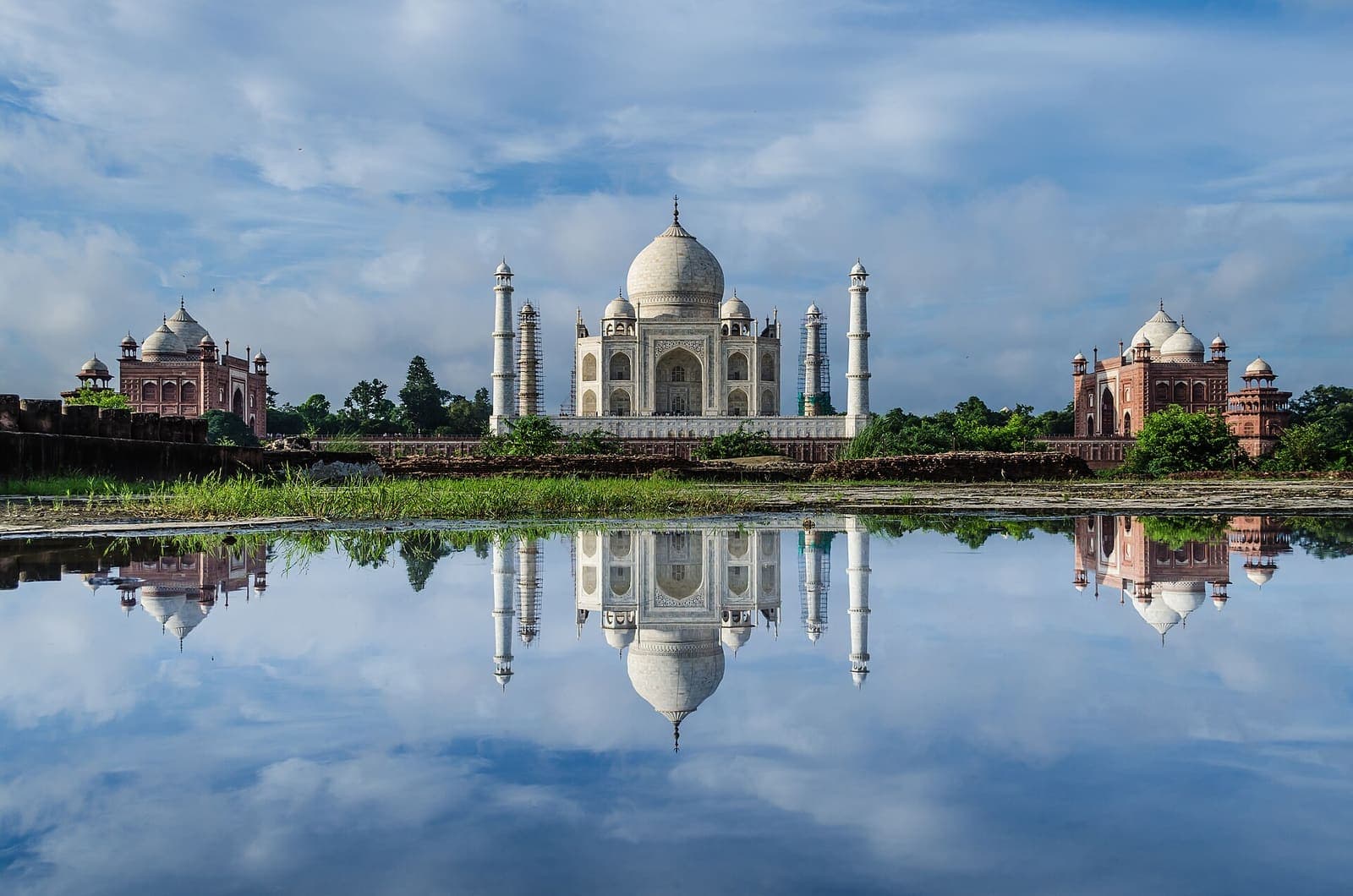 The Taj Mahal across the Yamuna from the Mehtab Bagh side, mirrored in still water under a cloudy blue sky